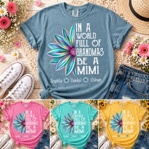 Könnte beinhalten: Ein hellblaues T-Shirt mit dem Text "IN A WORLD FULL OF GRANDMAS BE A MIMI" und einem bunten Blumendesign. Die Namen Sophia, Daniel und Ethan sind darunter aufgedruckt. Zusätzliche Shirts in Pink, Türkis und Gelb sind ebenfalls abgebildet.