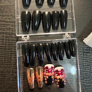 May include: A set of black, coffin-shaped press-on nails in a clear plastic case. Some nails are plain black, while others feature gold and red accents with decorative embellishments. A few nails are a metallic gold color.
