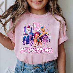 May include: Pink t-shirt featuring a colorful graphic of animated ponies, the number 4, and the name "Audrianna" in a playful font. The shirt is a short-sleeved design.