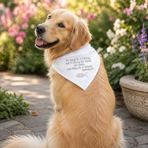 May include: A golden retriever dog wearing a white bandana with Greek text and a fish symbol. The dog is sitting outdoors with a blurred background of pink and white flowers. The dog is looking to the side with its tongue out.
