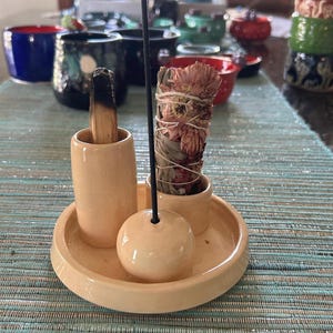 May include: A peach-colored ceramic incense holder set featuring a circular tray, a tall cylindrical holder for a palo santo stick, a small holder for a sage bundle with dried pink flowers, and a central sphere for a thin black incense stick.