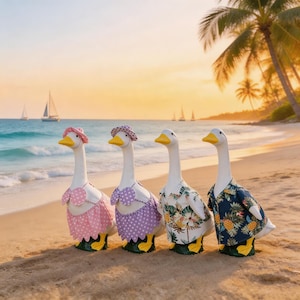 Puede incluir: Cuatro figuras de gansos blancos en una playa de arena, cada uno con diferentes atuendos. Dos gansos visten trajes de baño y sombreros, mientras que los otros dos lucen camisas hawaianas. El fondo presenta una puesta de sol, el océano y veleros.