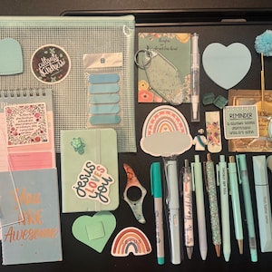 May include: An assortment of stationery and accessories in shades of mint green and pastel colors. Includes pens, a notebook, stickers, a planner, a clear pouch, and decorative items. The text "You Are Awesome" is visible on a small notebook.