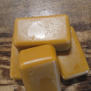 May include: Four rectangular bars of golden-yellow wax are stacked on a wooden surface. The wax has a smooth, slightly reflective surface with some areas appearing lighter in color. The bars are arranged in a slightly overlapping pattern.