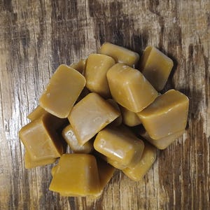 May include: A pile of golden-brown, square-shaped wax melts. The wax cubes have a smooth, glossy surface and are arranged on a rustic, wooden background. The color of the wax is a warm, inviting shade, suggesting a natural product.
