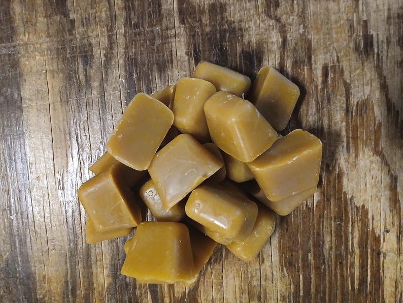 May include: A pile of golden-brown, square-shaped wax melts. The wax cubes have a smooth, glossy surface and are arranged on a rustic, wooden background. The color of the wax is a warm, inviting shade, suggesting a natural product.