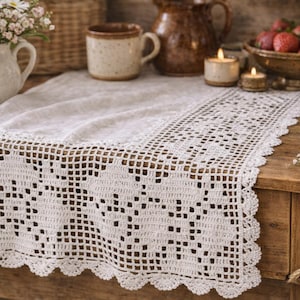 May include: A cream-colored, crocheted table runner with a scalloped edge. The runner is placed on a wooden table, with a vase of daisies, mugs, candles, and a bowl of strawberries in the background. The runner's design features openwork squares and decorative patterns.