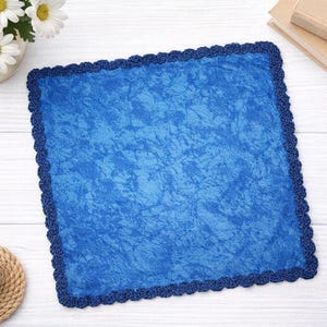 Könnte beinhalten: Ein quadratischer, blauer Stoff mit strukturiertem Aussehen und einem dunkelblauen Häkelrand. Der Stoff ist von einer dunkelblauen Häkelborte umgeben. Eine Vase mit weißen Gänseblümchen und eine Kerze auf einem Untersetzer sind ebenfalls zu sehen.