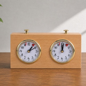 Puede incluir: Un reloj de ajedrez de madera con dos esferas redondas. Cada esfera tiene un fondo blanco, números negros y un borde dorado. Las manecillas son negras y el reloj está sobre una superficie de madera.