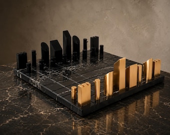 Può includere: Un moderno set di scacchi con una combinazione di colori nero e oro. La scacchiera è in marmo nero con un motivo a griglia. I pezzi degli scacchi sono neri e dorati, con forme geometriche. Il set è esposto su una superficie di marmo.