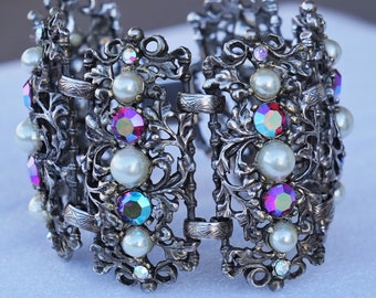 May include: Silver-toned filigree bracelet featuring faux pearls and iridescent, multi-colored rhinestones. The hinged bracelet has an ornate, openwork design with a clasp closure. A vintage-inspired piece of jewelry.
