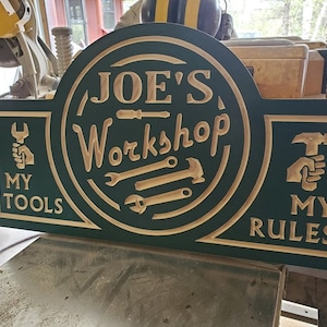 May include: A green wooden sign with the text "JOE'S Workshop" in a circle with tools around it. The sign also has the text "MY TOOLS" on the left and "MY RULES" on the right.