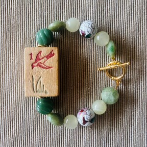 May include: A handmade bracelet featuring a wooden tile with a red bird design and the number 1. The bracelet includes green and white beads, and two decorative floral beads. It is secured with a gold-colored toggle clasp.