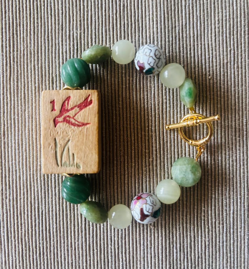 May include: A handmade bracelet featuring a wooden tile with a red bird design and the number 1. The bracelet includes green and white beads, and two decorative floral beads. It is secured with a gold-colored toggle clasp.