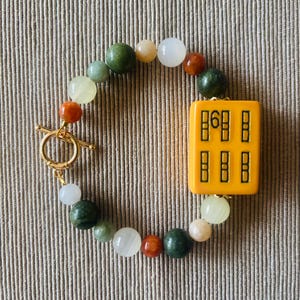 May include: A beaded bracelet featuring a gold toggle clasp and a central yellow rectangular charm with black symbols. The bracelet includes a mix of colorful round beads in shades of green, white, orange, and tan.