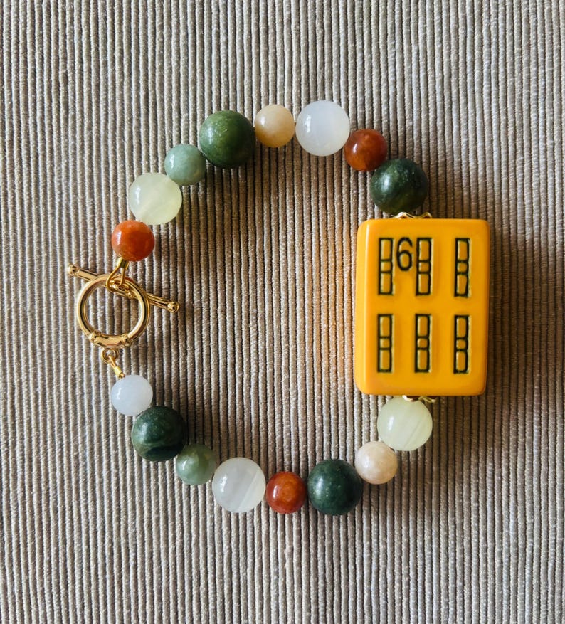 May include: A beaded bracelet featuring a gold toggle clasp and a central yellow rectangular charm with black symbols. The bracelet includes a mix of colorful round beads in shades of green, white, orange, and tan.