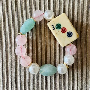 Puede incluir: Una pulsera de cuentas con una ficha de Mahjong rectangular de color blanco roto con el n&uacute;mero 3 y tres c&iacute;rculos de colores, intercalada con cuentas redondas rosa p&aacute;lido, cuentas blancas nacaradas y cuentas ovaladas verde claro.