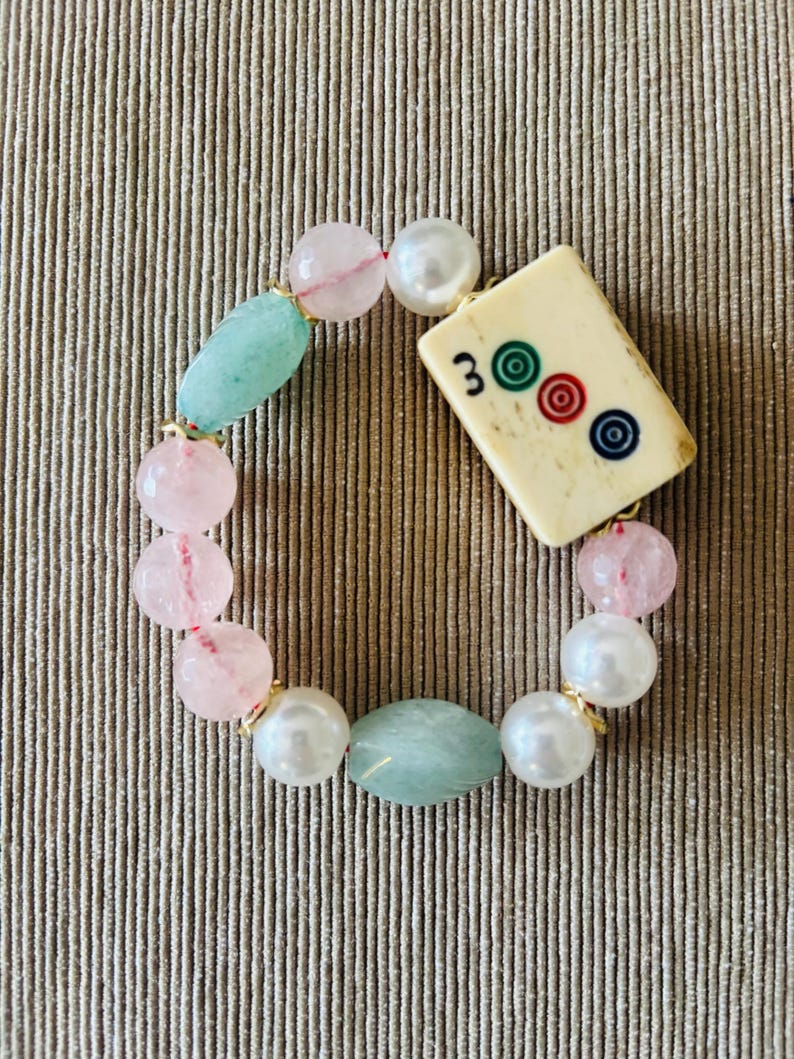 Puede incluir: Una pulsera de cuentas con una ficha de Mahjong rectangular de color blanco roto con el n&uacute;mero 3 y tres c&iacute;rculos de colores, intercalada con cuentas redondas rosa p&aacute;lido, cuentas blancas nacaradas y cuentas ovaladas verde claro.