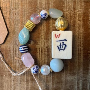 Puede incluir: Una pulsera de cuentas con una ficha de mahjong rectangular de color blanco roto con una letra W roja y un car&aacute;cter chino azul. La pulsera incluye cuentas de color rosa claro, azul p&aacute;lido y blanco, cuentas florales, rayadas y detalles dorados.