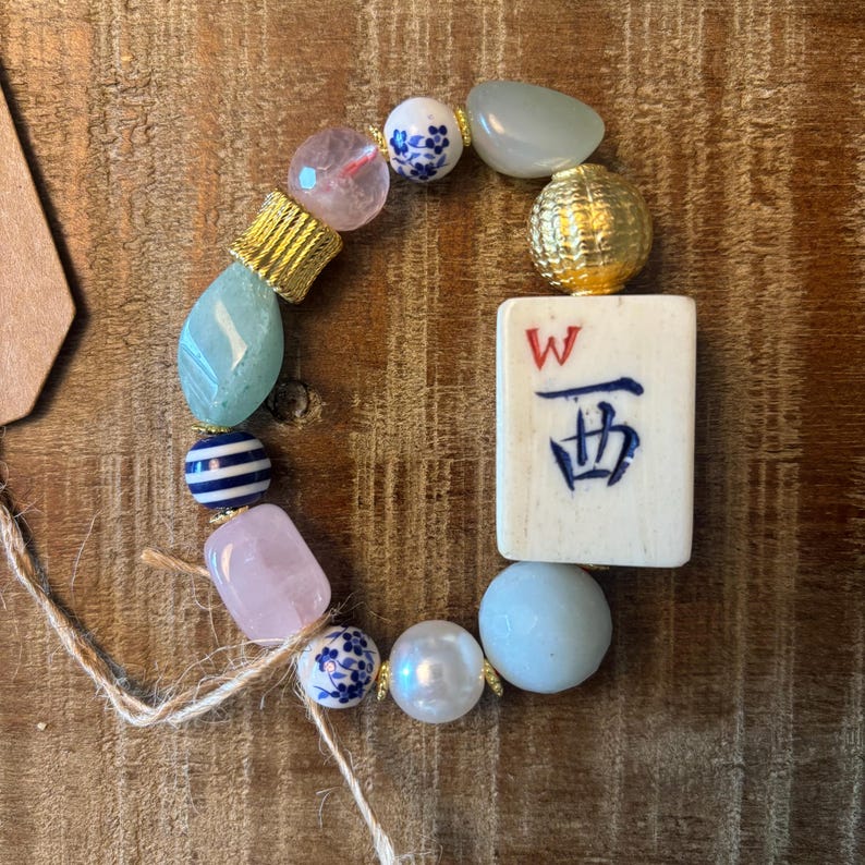 Puede incluir: Una pulsera de cuentas con una ficha de mahjong rectangular de color blanco roto con una letra W roja y un car&aacute;cter chino azul. La pulsera incluye cuentas de color rosa claro, azul p&aacute;lido y blanco, cuentas florales, rayadas y detalles dorados.