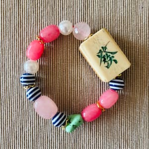 Puede incluir: Una pulsera de cuentas con una ficha de mahjong rectangular de color blanco roto con un car&aacute;cter verde, intercalada con cuentas lisas rosas, rosa claro y verdes, perlas blancas y peque&ntilde;as cuentas esf&eacute;ricas a rayas azules y blancas.