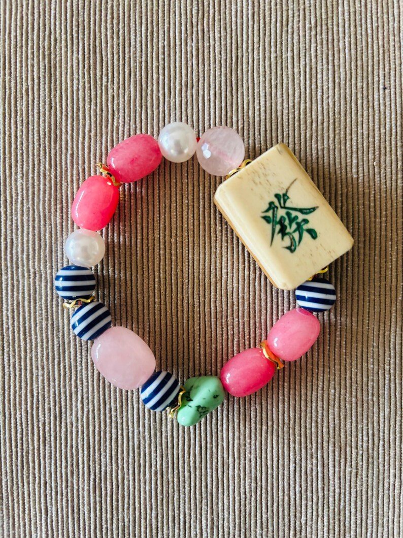 Puede incluir: Una pulsera de cuentas con una ficha de mahjong rectangular de color blanco roto con un car&aacute;cter verde, intercalada con cuentas lisas rosas, rosa claro y verdes, perlas blancas y peque&ntilde;as cuentas esf&eacute;ricas a rayas azules y blancas.