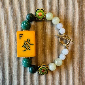 May include: A beaded bracelet featuring a yellow Mahjong tile with Chinese characters and the letter "F". The bracelet includes green and white beads, gold-colored flower accents, and a gold toggle clasp.