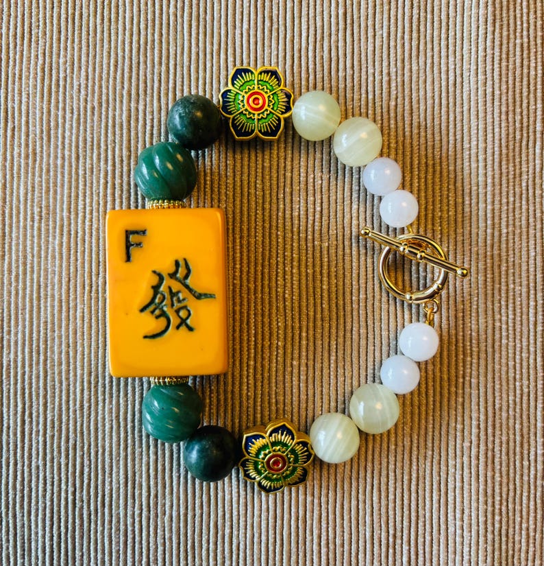 May include: A beaded bracelet featuring a yellow Mahjong tile with Chinese characters and the letter "F". The bracelet includes green and white beads, gold-colored flower accents, and a gold toggle clasp.