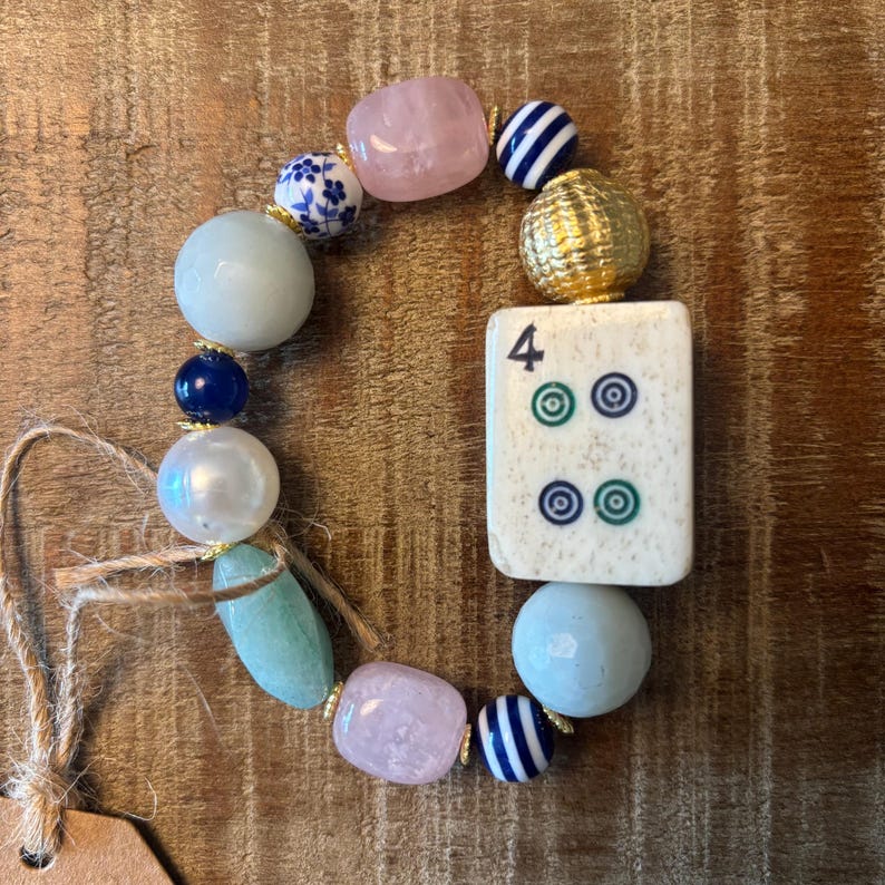 Puede incluir: Una pulsera de cuentas con una ficha de mahjong rectangular blanca con el n&uacute;mero 4 y s&iacute;mbolos circulares verdes y azules. La pulsera incluye cuarzo rosa, cuentas azules y blancas con motivos florales, cuentas azul marino, cuentas a rayas, una cuenta dorada texturizada y piedras azul claro.
