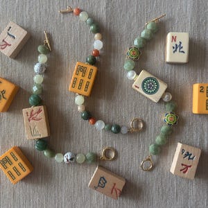 May include: Two beaded bracelets with green, white, and orange beads, and gold clasps. The bracelets feature decorative Mahjong tiles. Several Mahjong tiles are scattered around the bracelets on a neutral surface.