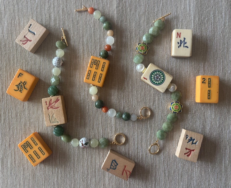 May include: Two beaded bracelets with green, white, and orange beads, and gold clasps. The bracelets feature decorative Mahjong tiles. Several Mahjong tiles are scattered around the bracelets on a neutral surface.