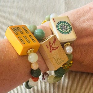 May include: A beaded bracelet featuring vintage Mahjong tiles in yellow and wood tones, accented with green and white beads. The bracelet is worn on a wrist, showcasing the colorful design and unique charm.