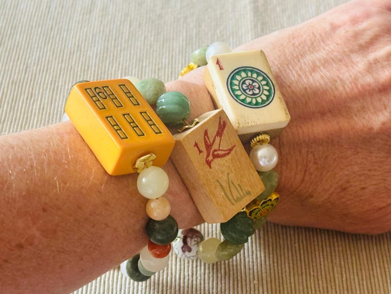 May include: A beaded bracelet featuring vintage Mahjong tiles in yellow and wood tones, accented with green and white beads. The bracelet is worn on a wrist, showcasing the colorful design and unique charm.