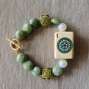 May include: A beaded bracelet featuring green gemstone beads, a gold toggle clasp, and decorative gold flower accents. The bracelet includes a Mahjong tile with the number 1 and a floral design.