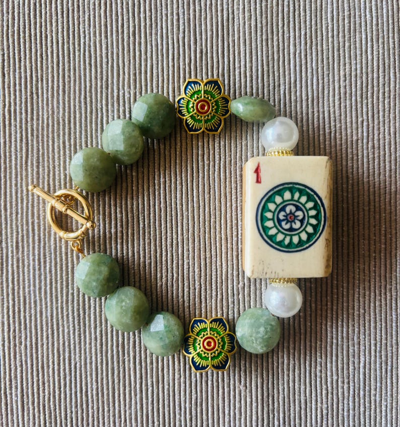 May include: A beaded bracelet featuring green gemstone beads, a gold toggle clasp, and decorative gold flower accents. The bracelet includes a Mahjong tile with the number 1 and a floral design.