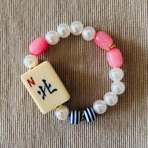 Puede incluir: Una pulsera de cuentas con una ficha de Mahjong rectangular de color blanco roto con la letra roja N y caracteres chinos negros. La banda consta de perlas blancas artificiales, cuentas ovaladas rosas lisas y dos cuentas esf&eacute;ricas a rayas azules y blancas.