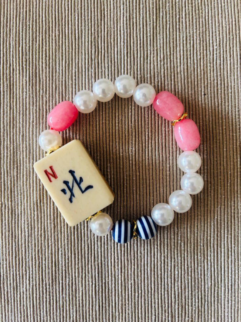 Puede incluir: Una pulsera de cuentas con una ficha de Mahjong rectangular de color blanco roto con la letra roja N y caracteres chinos negros. La banda consta de perlas blancas artificiales, cuentas ovaladas rosas lisas y dos cuentas esf&eacute;ricas a rayas azules y blancas.
