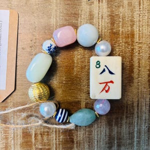 Puede incluir: Una pulsera de cuentas que presenta una ficha de mahjong rectangular con el n&uacute;mero 8 y caracteres chinos, intercalada con varias cuentas que incluyen un cuadrado rosa, esferas blancas, una bola dorada texturizada, cuentas a rayas y piedras verdes.