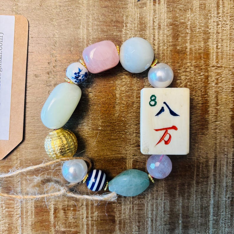 Puede incluir: Una pulsera de cuentas que presenta una ficha de mahjong rectangular con el n&uacute;mero 8 y caracteres chinos, intercalada con varias cuentas que incluyen un cuadrado rosa, esferas blancas, una bola dorada texturizada, cuentas a rayas y piedras verdes.