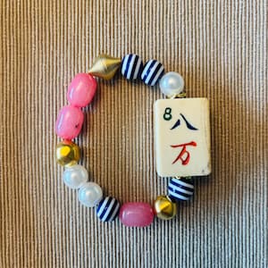 Puede incluir: Una pulsera de cuentas con una ficha de mahjong rectangular de color blanco roto con el n&uacute;mero 8 y caracteres chinos en verde, azul y rojo. La pulsera incluye cuentas rosas lisas, perlas blancas, cuentas met&aacute;licas doradas y cuentas a rayas azules y blancas.