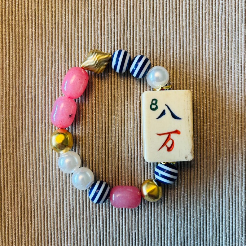 Puede incluir: Una pulsera de cuentas con una ficha de mahjong rectangular de color blanco roto con el n&uacute;mero 8 y caracteres chinos en verde, azul y rojo. La pulsera incluye cuentas rosas lisas, perlas blancas, cuentas met&aacute;licas doradas y cuentas a rayas azules y blancas.