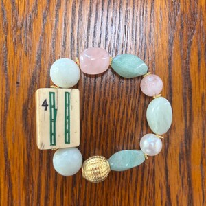Puede incluir: Una pulsera el&aacute;stica que presenta una ficha de mahjong rectangular de color blanco hueso con un n&uacute;mero cuatro verde y un dise&ntilde;o de bamb&uacute;, intercalada con cuentas lisas redondas y ovaladas en tonos rosa p&aacute;lido, verde menta y blanco, y una cuenta dorada.