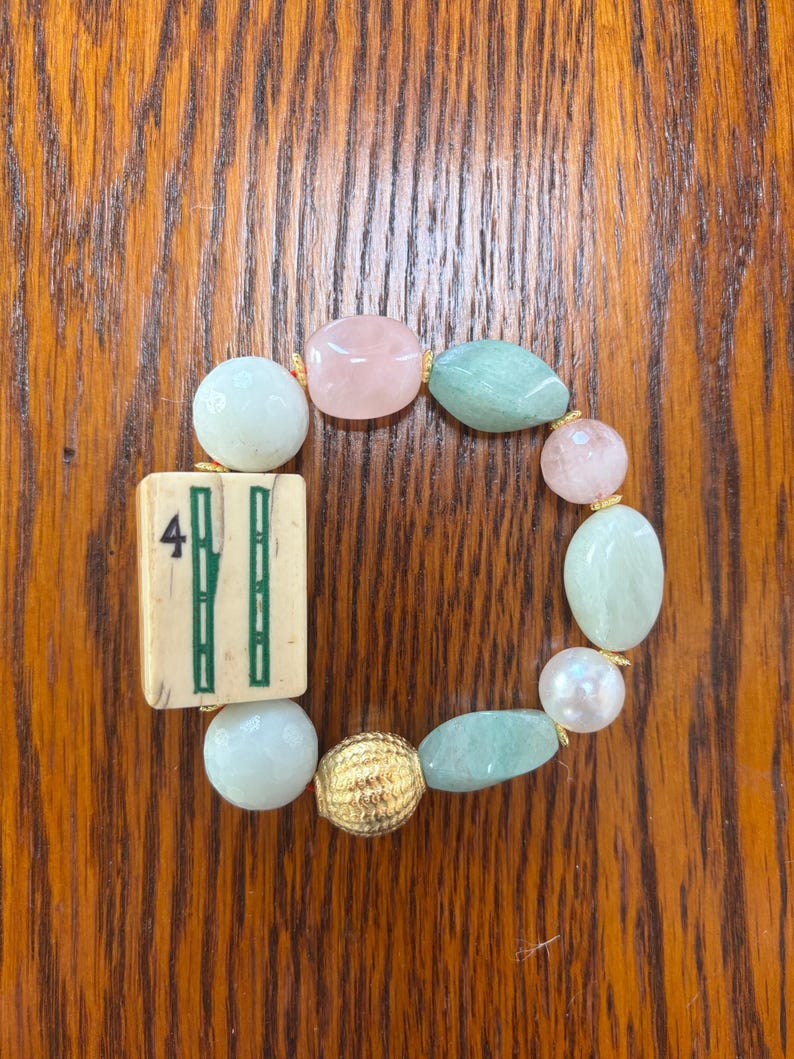 Puede incluir: Una pulsera el&aacute;stica que presenta una ficha de mahjong rectangular de color blanco hueso con un n&uacute;mero cuatro verde y un dise&ntilde;o de bamb&uacute;, intercalada con cuentas lisas redondas y ovaladas en tonos rosa p&aacute;lido, verde menta y blanco, y una cuenta dorada.