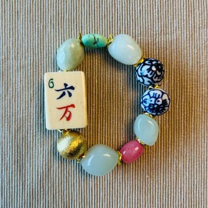 Puede incluir: Una pulsera el&aacute;stica que presenta una ficha de mahjong rectangular con el n&uacute;mero 6 y caracteres chinos en azul y rojo. La pulsera incluye cuentas lisas de color verde claro, dos cuentas de porcelana azul y blanca, una cuenta rosa y una cuenta dorada.