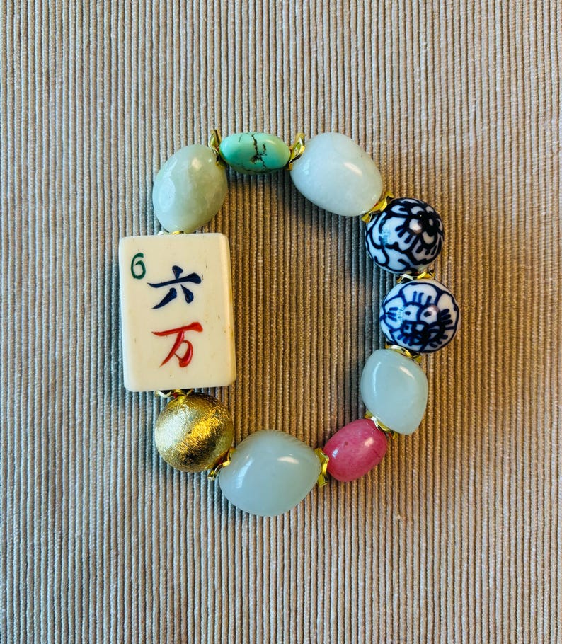 Puede incluir: Una pulsera el&aacute;stica que presenta una ficha de mahjong rectangular con el n&uacute;mero 6 y caracteres chinos en azul y rojo. La pulsera incluye cuentas lisas de color verde claro, dos cuentas de porcelana azul y blanca, una cuenta rosa y una cuenta dorada.