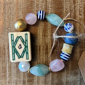 Puede incluir: Una pulsera de cuentas con una ficha de mahjong rectangular con marcas de bamb&uacute; verdes, intercalada con cuarzo rosa, cuentas a rayas azules y blancas, aventurina verde, una cuenta dorada en forma de rosa, una cuenta azul floral y un espaciador dorado.