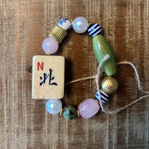 Puede incluir: Una pulsera de cuentas con una ficha de mahjong color crema con la letra N y un car&aacute;cter negro, intercalada con cuarzo rosa, perlas blancas, cuentas a rayas azules y blancas, una cuenta cil&iacute;ndrica verde y una cuenta dorada con dise&ntilde;o floral.