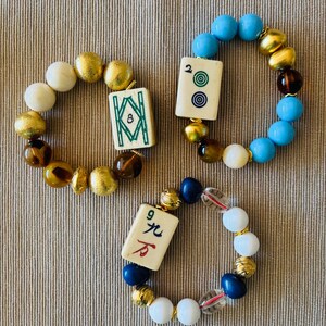 Puede incluir: Tres pulseras el&aacute;sticas con fichas de mahjong y cuentas variadas. Una pulsera tiene una ficha de bamb&uacute; 8 con cuentas doradas y marrones. Otra tiene una ficha de 2 puntos con cuentas azules, doradas y marrones. La tercera tiene una ficha de car&aacute;cter 9 con cuentas blancas, azules y doradas.