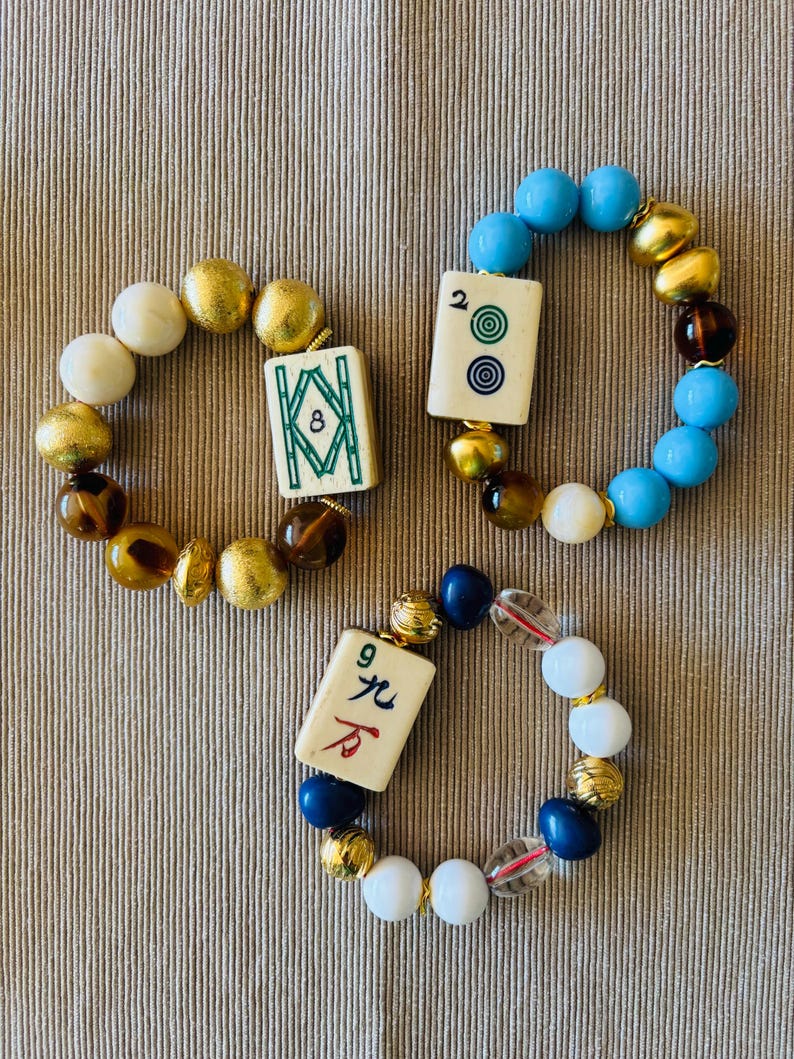 Puede incluir: Tres pulseras el&aacute;sticas con fichas de mahjong y cuentas variadas. Una pulsera tiene una ficha de bamb&uacute; 8 con cuentas doradas y marrones. Otra tiene una ficha de 2 puntos con cuentas azules, doradas y marrones. La tercera tiene una ficha de car&aacute;cter 9 con cuentas blancas, azules y doradas.