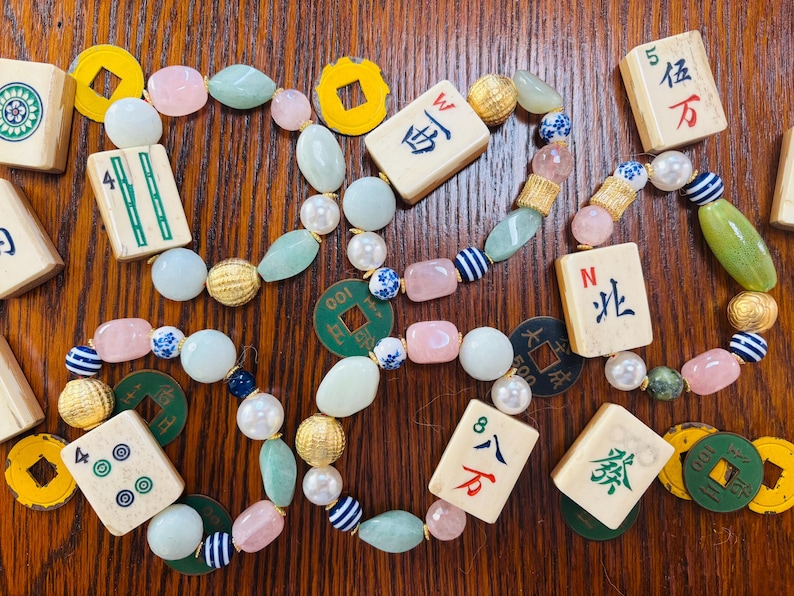 Puede incluir: Tres pulseras hechas con fichas de mahjong, cuentas de color verde claro tipo jade, cuentas de cuarzo rosa, perlas blancas, esferas doradas texturizadas y cuentas a rayas azules y blancas. Las fichas muestran caracteres chinos y bamb&uacute;es.